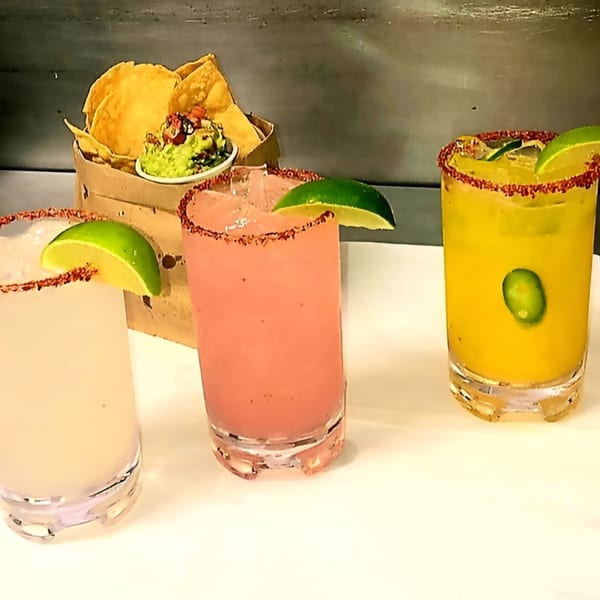 Three colorful Fuego Mexican Kitchen margaritas with lime wedges and a basket of chips and guacamole.