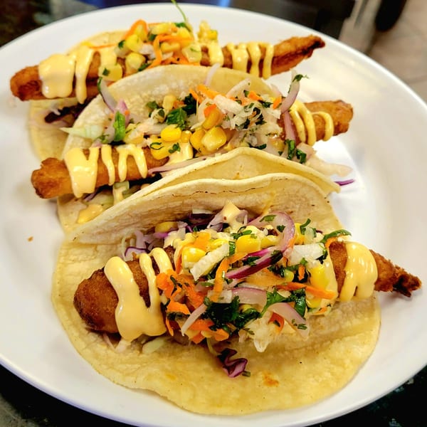 Three Fuego Mexican Kitchen fish tacos with corn, cabbage, and creamy sauce on a white plate.