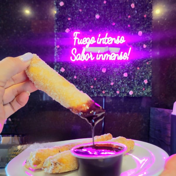 Hand dipping a churro into chocolate sauce, with a neon sign and green wall in the background.